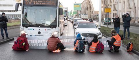 Auf einer Straße sitzen fünf Menschen auf dem Boden. Dahinter stauen sich Autos und ein Bus. Daneben sind Pressemenschen mit Kameras zu sehen.
