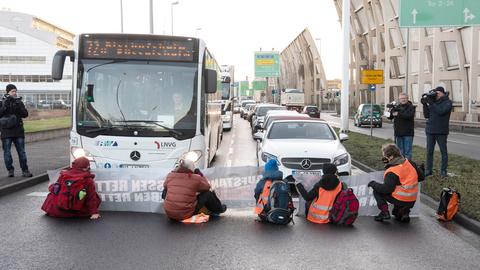 Auf einer Straße sitzen fünf Menschen auf dem Boden. Dahinter stauen sich Autos und ein Bus. Daneben sind Pressemenschen mit Kameras zu sehen.