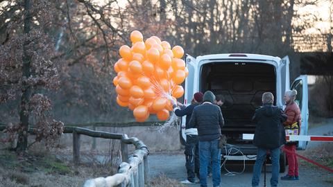 Ein Mann hält mehrere orange Luftballons an Schnüren in der Hand, daneben ein Transporter mit offenem Kofferraum und weitere Menschen.