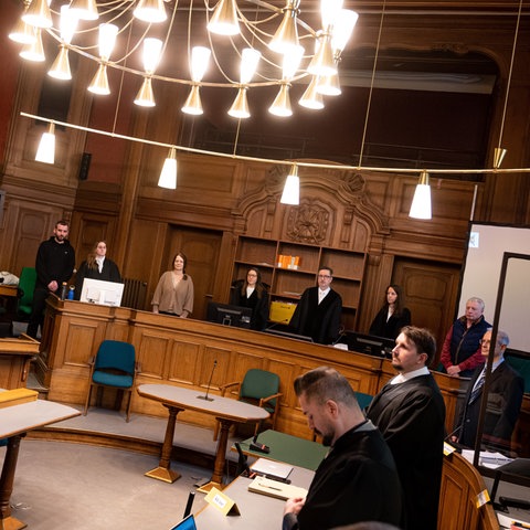 In einem Gerichtssaal stehen mehrere Menschen hinter Pulten.