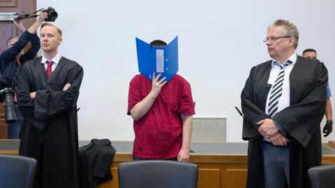 Angeklagter in T-Shirt im Gerichtssaal - er hält sich einen geöffneten Ordner vor das Gesicht. Rechts und links daneben Anwälte, ein Reporter mit Kamera und Polizist
