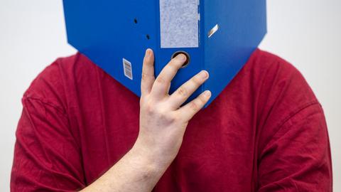 Mann in rotem Shirt, der sich einen blauen Aktenordner vors Gesicht hält.