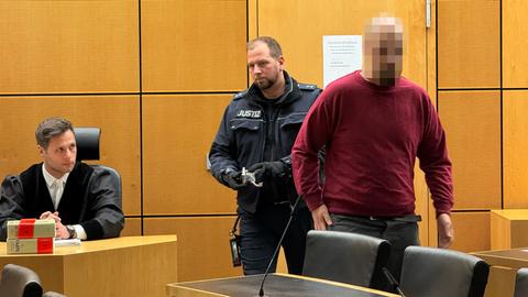 Ein Mann in rotem Pullover wird von einem Justizbeamten in einen Gerichtssaal geführt.