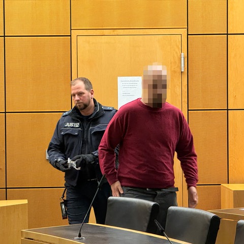 Ein Mann in rotem Pullover wird von einem Justizbeamten in einen Gerichtssaal geführt.