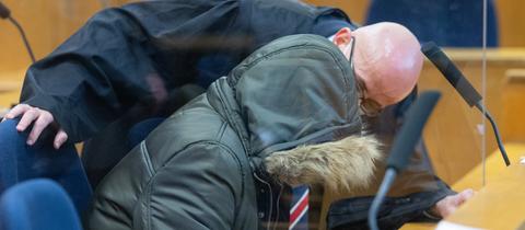 Der Angeklagte trägt eine Kapuzenjacke, die sein Gesicht verdeckt. Daneben beugt sich sein Anwalt zu ihm herunter, um mit ihm zu sprechen.