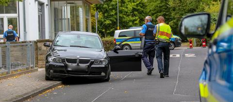 Ein schwarzer BMW steht mit geöffneter Fahrertür teilweise auf einem Bürgersteig und der Straße. Polizisten gehen daran vorbei. 