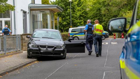 Ein schwarzer BMW steht mit geöffneter Fahrertür teilweise auf einem Bürgersteig und der Straße. Polizisten gehen daran vorbei. 