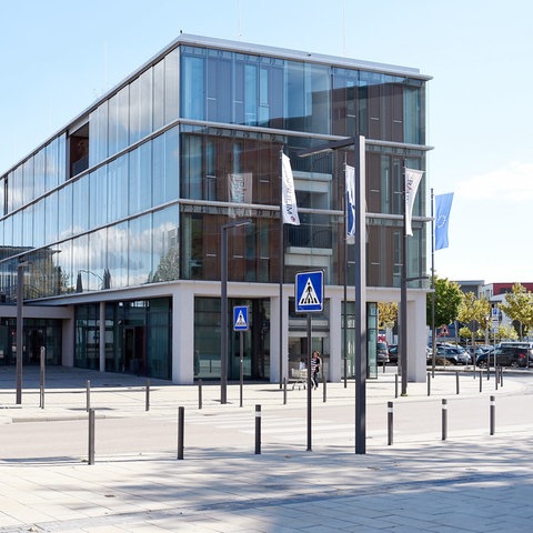 Zeitgenössiches Gebäude mit Glasfassade, auf einem Platz mit Straße und Bushaltestelle stehend.
