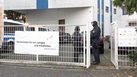 Vor einem Gebäude stehen zwei maskierte und bewaffnete Einsatzkräfte der Polizei. Am Zaun des Geländes hängt ein Banner mit der Aufschrift "Gemeinsam für ein ruhiges und friedliches Rödelheim" und einer Friedenstaube