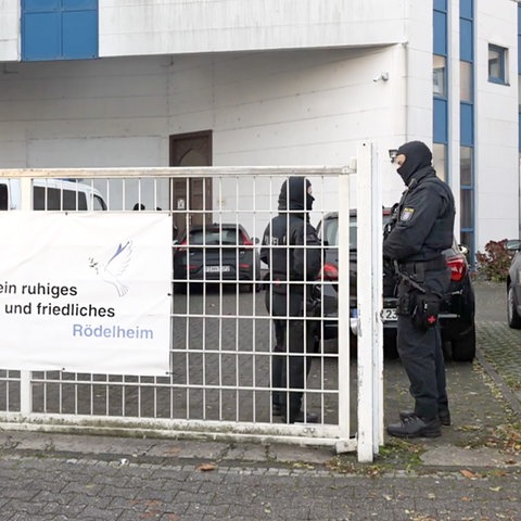 Vor einem Gebäude stehen zwei maskierte und bewaffnete Einsatzkräfte der Polizei. Am Zaun des Geländes hängt ein Banner mit der Aufschrift "Gemeinsam für ein ruhiges und friedliches Rödelheim" und einer Friedenstaube