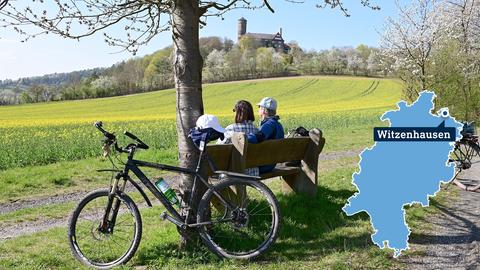Foto: Zwei Radfahrer machen Rast auf einer Bank (im Bildvordergrund). In der Ferne (Bildhintergrund) ist die Burg Ludwigstein auf einer Bergkuppe zu sehen. Auf dem Feld vor ihnen fängt der Raps zartgelb an zu blühen. Im Foto ist rechts eine kleine, blaue Hessenkarte mit der Stadt Witzenhausen verortet. 