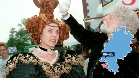 Foto: Eine Frau und ein Mann kostümiert in Biedermeier-Kleidung. Im Foto ist rechts eine kleine, blaue Hessenkarte mit dem Ort Eltville. 