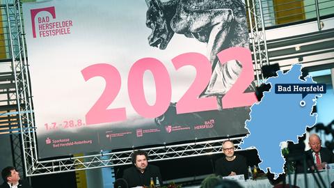 Foto: Vier Männer sitzen an Tischen. Über ihnen ein riesiges Plakat, auf welchem "Bad Hersfelder Festspiele  - 2022" in rosa Lettern steht. Im Foto ist rechts eine kleine, blaue Hessenkarte mit dem Ort Bad Hersfeld. 