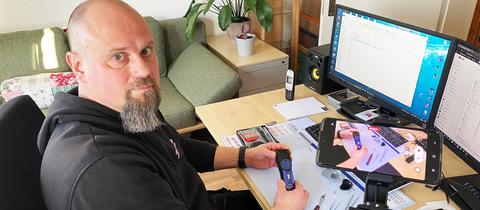 Stephan Heinrich sitzt am Schreibtisch und hat einen Computer, ein Smartphone im Aufnahmemodus und ein technisches Gerät vor und neben sich. Er blickt in die Kamera.