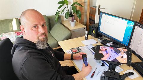 Stephan Heinrich sitzt am Schreibtisch und hat einen Computer, ein Smartphone im Aufnahmemodus und ein technisches Gerät vor und neben sich. Er blickt in die Kamera.