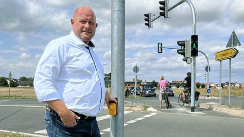 Marcus Kretschmann steht an der Ampelkreuzung mit der Hand am Knopf für die Fußgänger:innen. Im Hintergund eine Kreuzung mit Ampel und Passanten.