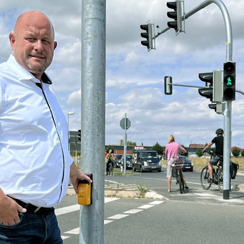 Marcus Kretschmann steht an der Ampelkreuzung mit der Hand am Knopf für die Fußgänger:innen. Im Hintergund eine Kreuzung mit Ampel und Passanten.