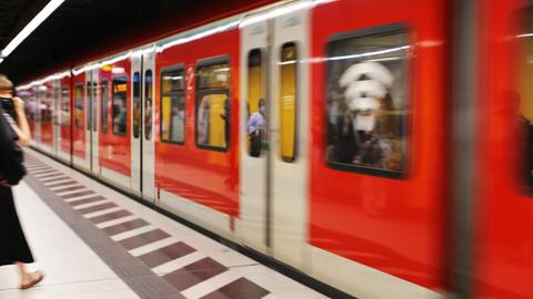 An einem Bahnsteig fährt eine S-Bahn - unscharf - ein. In den Fenster spiegeln sich wartende Personen.