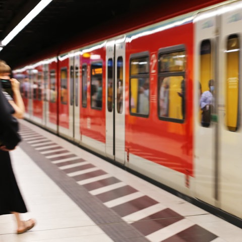 An einem Bahnsteig fährt eine S-Bahn - unscharf - ein. In den Fenster spiegeln sich wartende Personen.