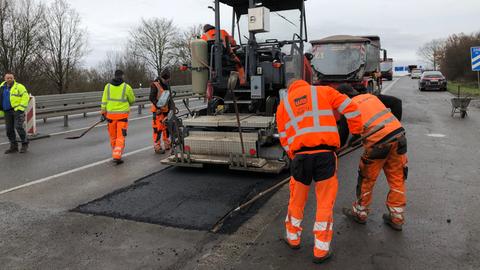 Um eine große Teermaschine (in der Mitte des Bildes) herum bringen Bauarbeiter in neonfarbenen Arbeitsanzügen ein neues schwarzes Teerfeld auf eine Straße auf. 