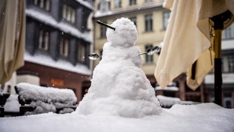 Ein kleiner Schneemann mit einer Nase aus einem Bleistift und Armen aus Löffeln steht auf dem Tisch im verschneiten Außenbereich eines Cafés in der neuen Frankfurter Altstadt.