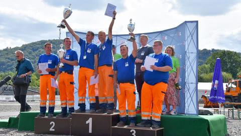 Mehrere Männer in blauen T-Shirts und orangen Hosen stehen mit Pokalen auf einem Siegerpodest.