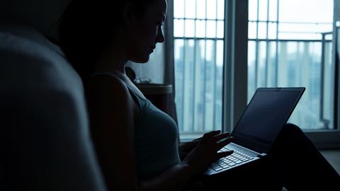 Eine Frau sitzt im Dunkel der Dämmerung vor einem Fenster und schreibt auf einem Laptop.