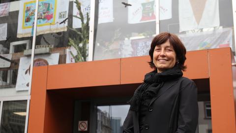 Eine Frau mit kurzen schwarzen Haaren und einem schwarzen Blazer steht vor einer Glasfassade.