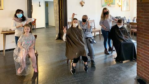 Im Innenraum einer alten Stube: Drei Frauen stehen hinter drei Menschen, die vor ihnen sitzen und schneiden ihnen die Haare.