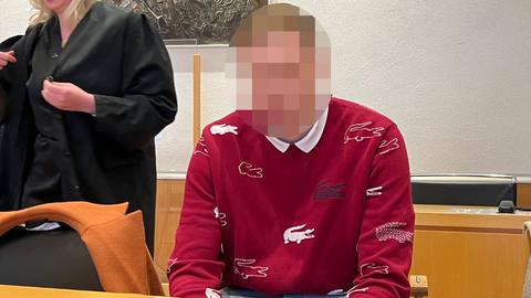 Ein Mann im roten Pulli sitzt an einem Tisch. Sein Kopf ist unkenntlich gemacht. Daneben eine Frau in schwarzer Robe.