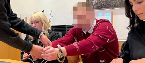 Ein Mann im roten Pulli sitzt an einem Tisch. Sein Kopf ist unkenntlich gemacht. Daneben eine Frau in schwarzer Robe.