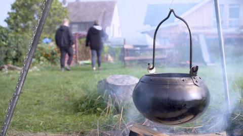 Über einem Feuer hängt ein Eisentopf, aus dem es köchelt. Im Hintergrund laufen zwei Menschen weg.