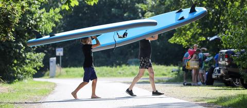Ein Vater und sein Sohn tragen ihre Stand-Up Paddling-Boards (SUPs) zu einem Badesee.