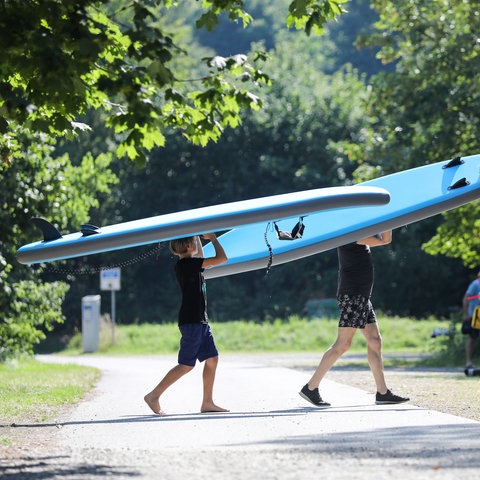 Ein Vater und sein Sohn tragen ihre Stand-Up Paddling-Boards (SUPs) zu einem Badesee.