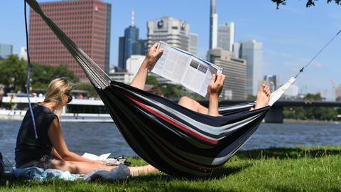 Eine Frau sitzt am Mainufer Frankfurt im Gras, ein Mann liegt in einer Hängematte, die daneben aufgespannt ist.