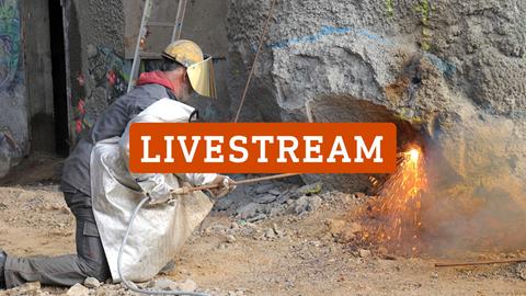 Ein Mann in schwerer Schutzkleidung bearbeitet mit Feuer und Funkenflug einen Pfleiler der Brücke. Auf dem Foto steht in großen Lettern "LIVE-STREAM".