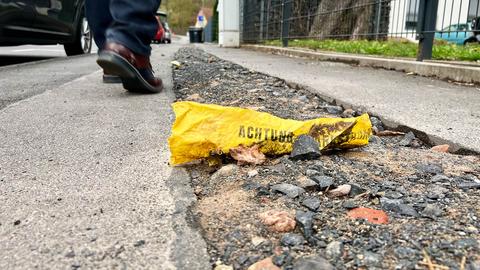 Nahaufnahme eines Bürgersteigs. Aus einer steinigen Fläche in der Mitte ragt ein gelbes zerknittertes Band mit der Aufschrift "Achtung". Dahinter sich bewegende Füße eines gehenden Menschen.