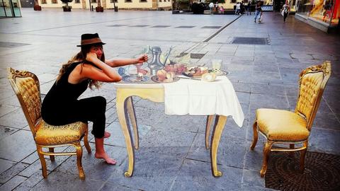 Eine Frau sitzt zwischen Häusern auf einem goldenen Stuhl neben einem Tisch, der als optische Täuschung auf das Pflaster gezeichnet wurde.