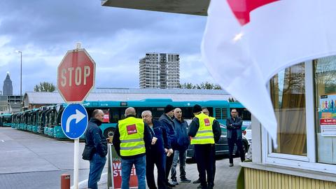 Busfahrer bekleidet mit Verdi-Warnwesten stehen vor neben einem kleinen Gebäude. Im Hintergund ein Parkplatz mit vielen Bussen, die geordnet nebeneinander stehen.