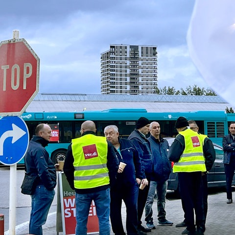 Busfahrer bekleidet mit Verdi-Warnwesten stehen vor neben einem kleinen Gebäude. Im Hintergund ein Parkplatz mit vielen Bussen, die geordnet nebeneinander stehen.