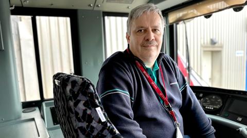 Das Bild zeigt einen älteren Mann mit grauen Haaren und Schnurrbart in der Fahrerkabine einer Straßenbahn. Er trägt einen dunkelblauen Pullover und eine Schlüsselkarte an einem Halsband. 