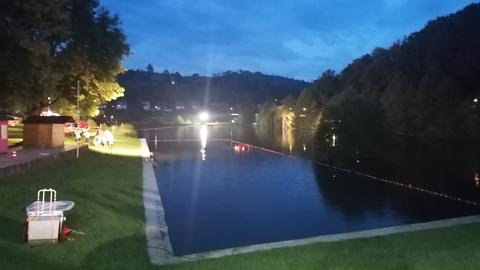 Ein See liegt im Dämmerlicht, im Hintergrund sind Einsatzkräfte der Feuerwehr zu sehen.