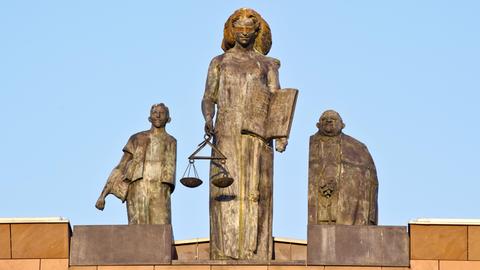 Steinerne Statue der Justitia in der Abendsonne auf einem Dach installiert. Sie wird eingerahmt von zwei weiteren Statuen.