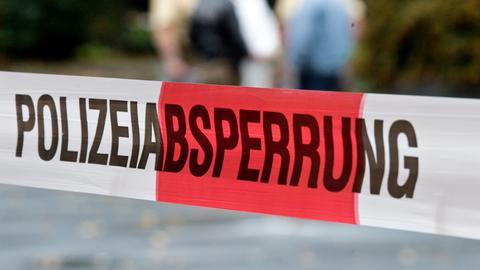 Absperrband, rot-weiß gestreift, mit der Aufschrift "Polizeiabsperrung". Im Bildhintergrund unscharf ein Grüppchen von Menschen.