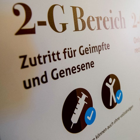 Ein Schild mit der Aufschrift "2-G Bereich, Zutritt für Geimpfte und Genesene". In Hintergrund ist unscharfn ein Weihnachtsmarkt zu sehen. 