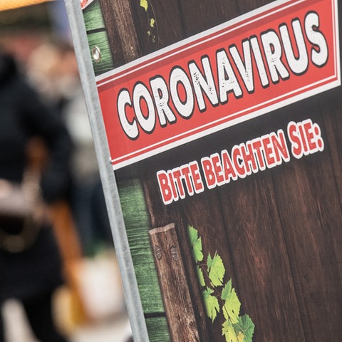 Schild mit der Aufschrift "Coronavirus - bitte beachten Sie". Im Bildhintergrund unscharf Menschen in einer Fußgängerzone.