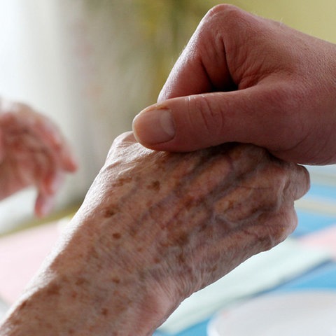 Großaufnahme: Eine jüngere Hand hält die Hand einer betagten Person.