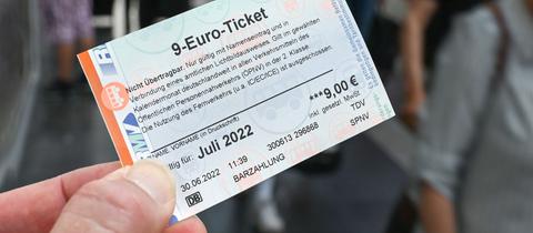 Eine Hand hält ein 9-Euro-Ticket (im Bildvordergrund scharf). Unscharf im Bildhintergrund Zugreisende am Bahnsteig.