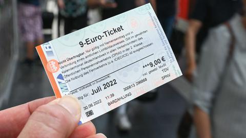 Eine Hand hält ein 9-Euro-Ticket (im Bildvordergrund scharf). Unscharf im Bildhintergrund Zugreisende am Bahnsteig.