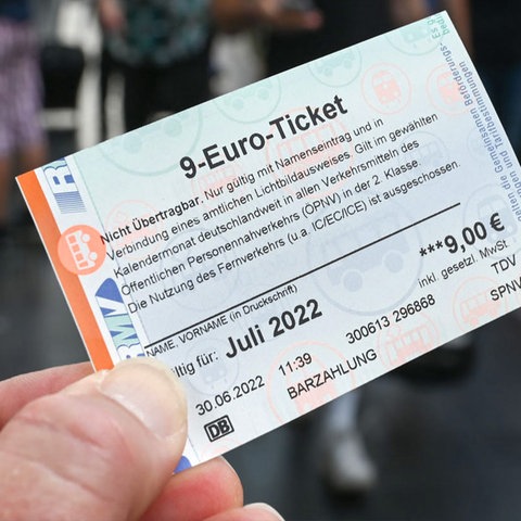 Eine Hand hält ein 9-Euro-Ticket (im Bildvordergrund scharf). Unscharf im Bildhintergrund Zugreisende am Bahnsteig.